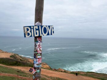 Big blue sign that says "BIG LOVE" in white letters is mounted on a wood pole on a bluff overlooking the ocean in Nazare, Portugal.
